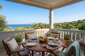 A round wooden table set for two with plates, glasses, and a bowl of fruit on a balcony overlooking a scenic landscape with trees and a view of the sea. - Villa Siesta  (Pangaio)