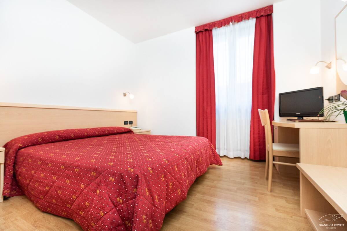 A hotel room with a bed, a desk, a chair, a TV, and a window with red curtains.