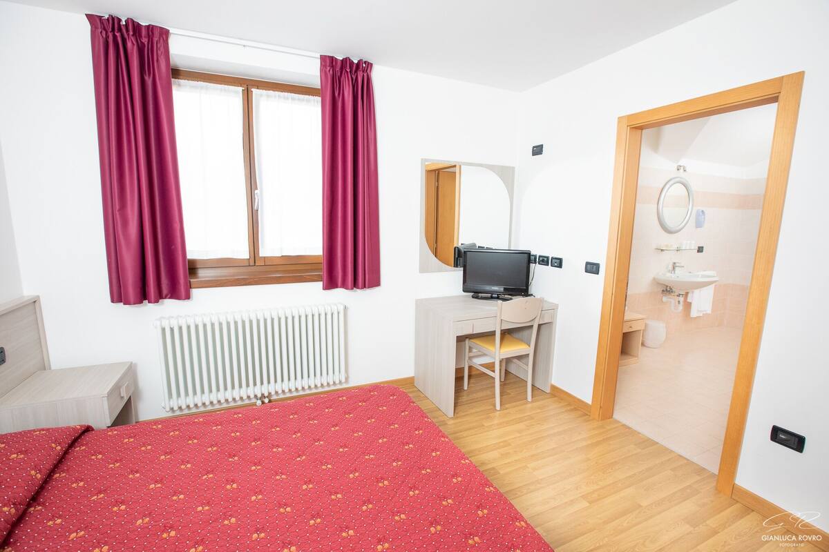 A bedroom with a bed, a desk, a chair, a computer, a window with curtains, a radiator, and a bathroom visible through an open door.