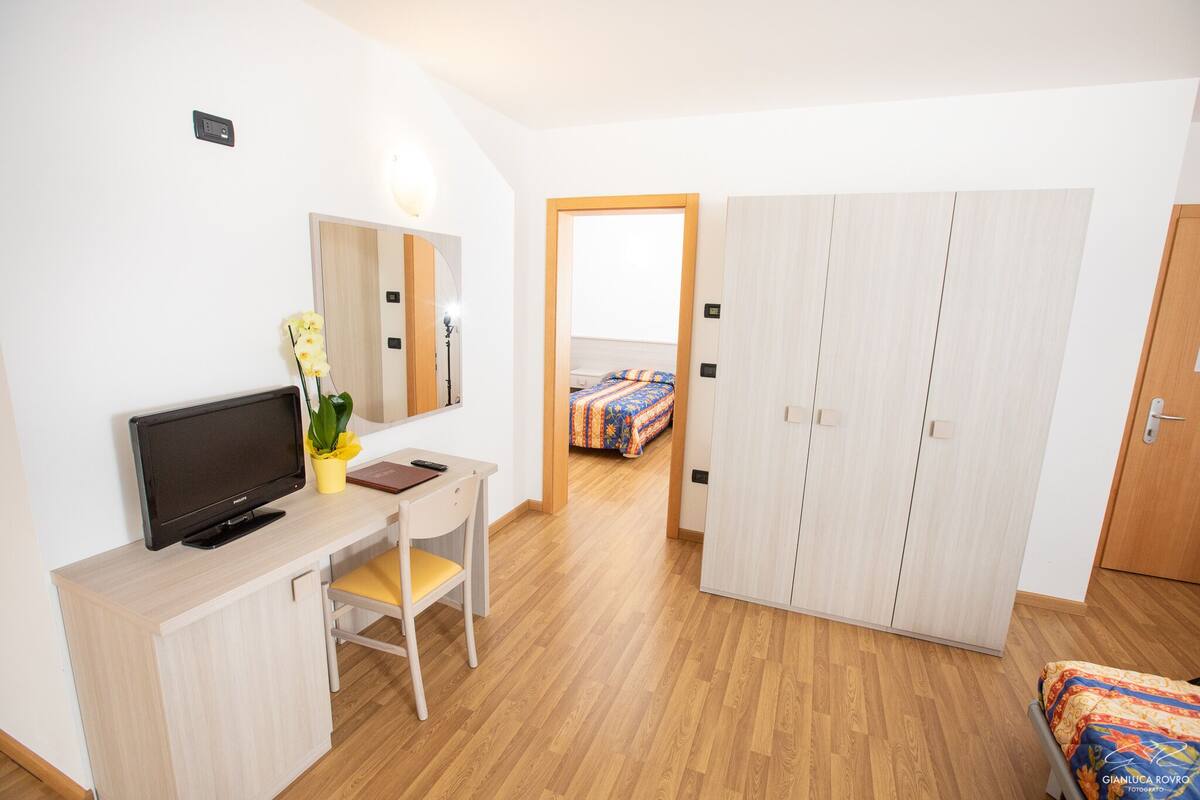 A modern hotel room with a wooden desk, a television, a chair, a wardrobe, and a bed with a colorful bedspread.