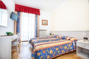 Un dormitori amb un llit, una taula de treball, una cadira, un televisor i una finestra amb cortines.