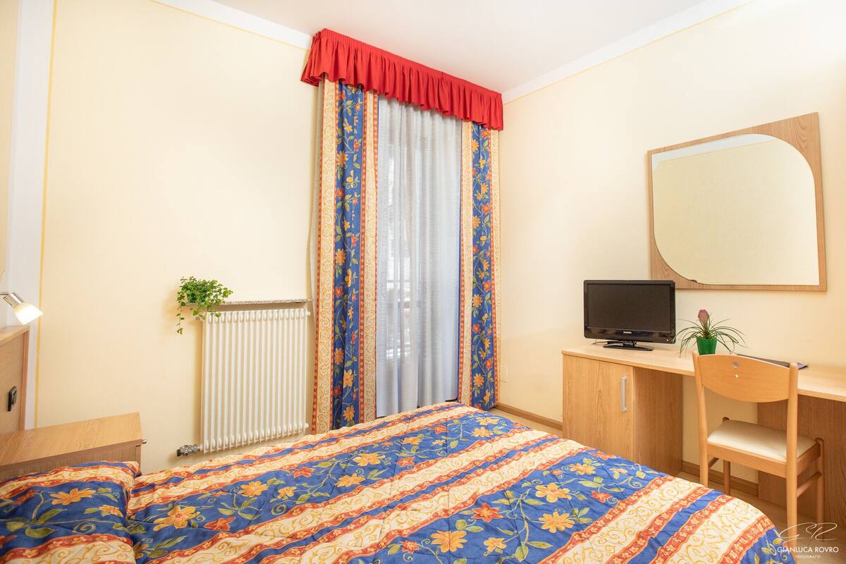 A bedroom with a bed, a desk with a computer, a chair, a radiator, and a window with curtains.