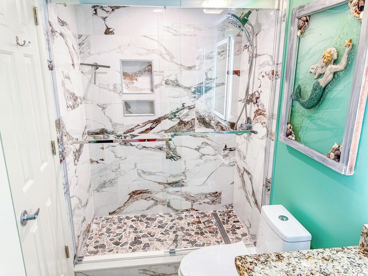 Een badkamer met een douchecabine van glas, marmeren tegels, een wastafel met een wit aanrechtblad en een decoratief muurschildering van een zeemeermin.