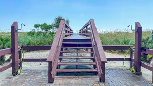 Un pont en bois doté d’une rambarde métallique, qui mène à une zone sablonneuse couverte de végétation.