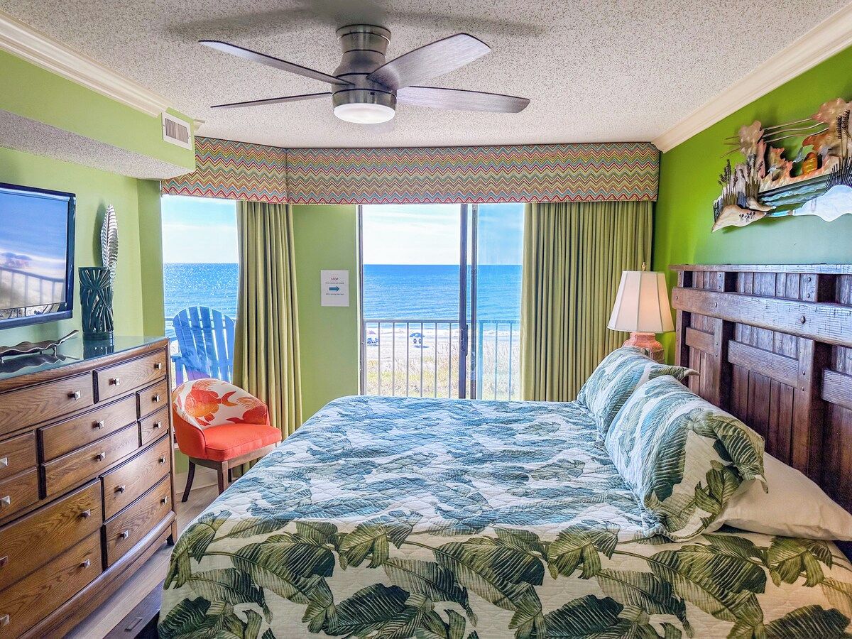 A bedroom with a bed, a dresser, a chair, a lamp, and a view of the sea.