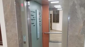 Un elevador moderno con puerta de vidrio y estructura metálica, que da a un pasillo con piso de baldosas y pared lisa.