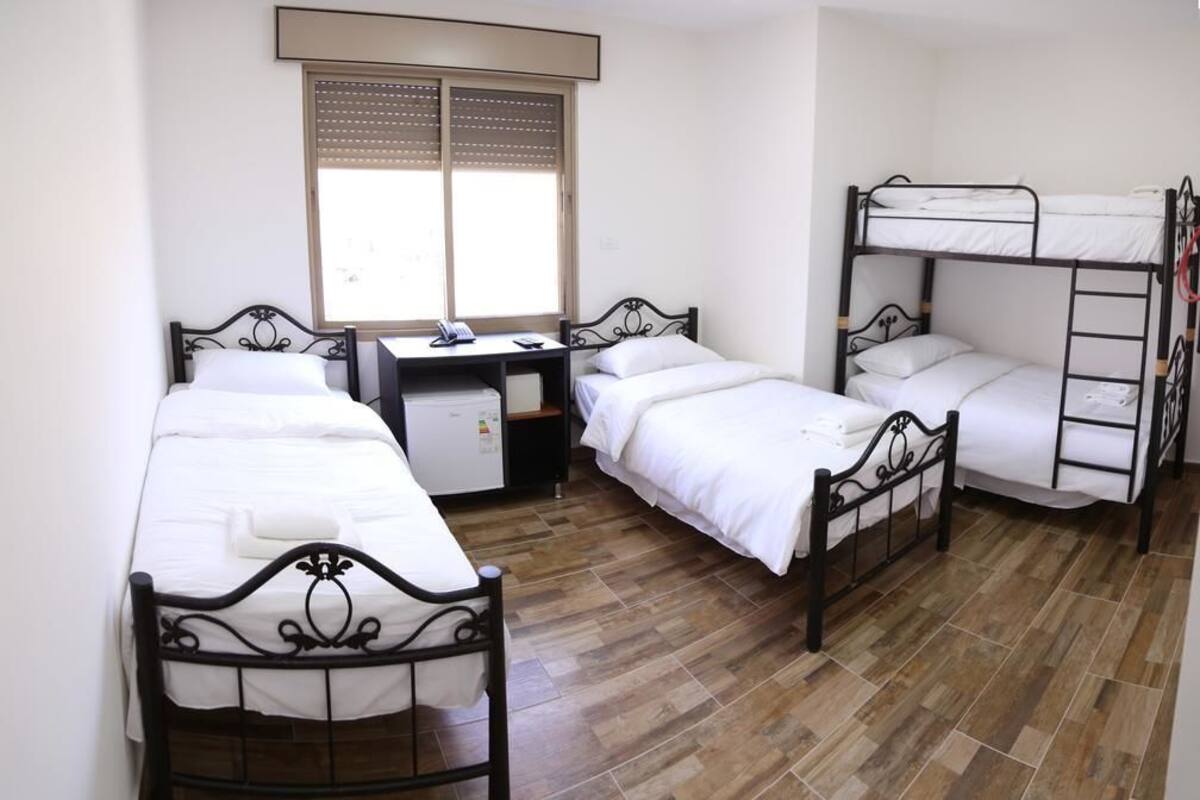 A dormitory room with three beds, a desk, and a window.