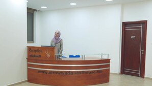 Un mostrador de recepción con terminación de madera y una superficie de vidrio, un cartel con el sitio web 'www.alrowwadguesthouse.com' y una puerta de baño a la derecha.