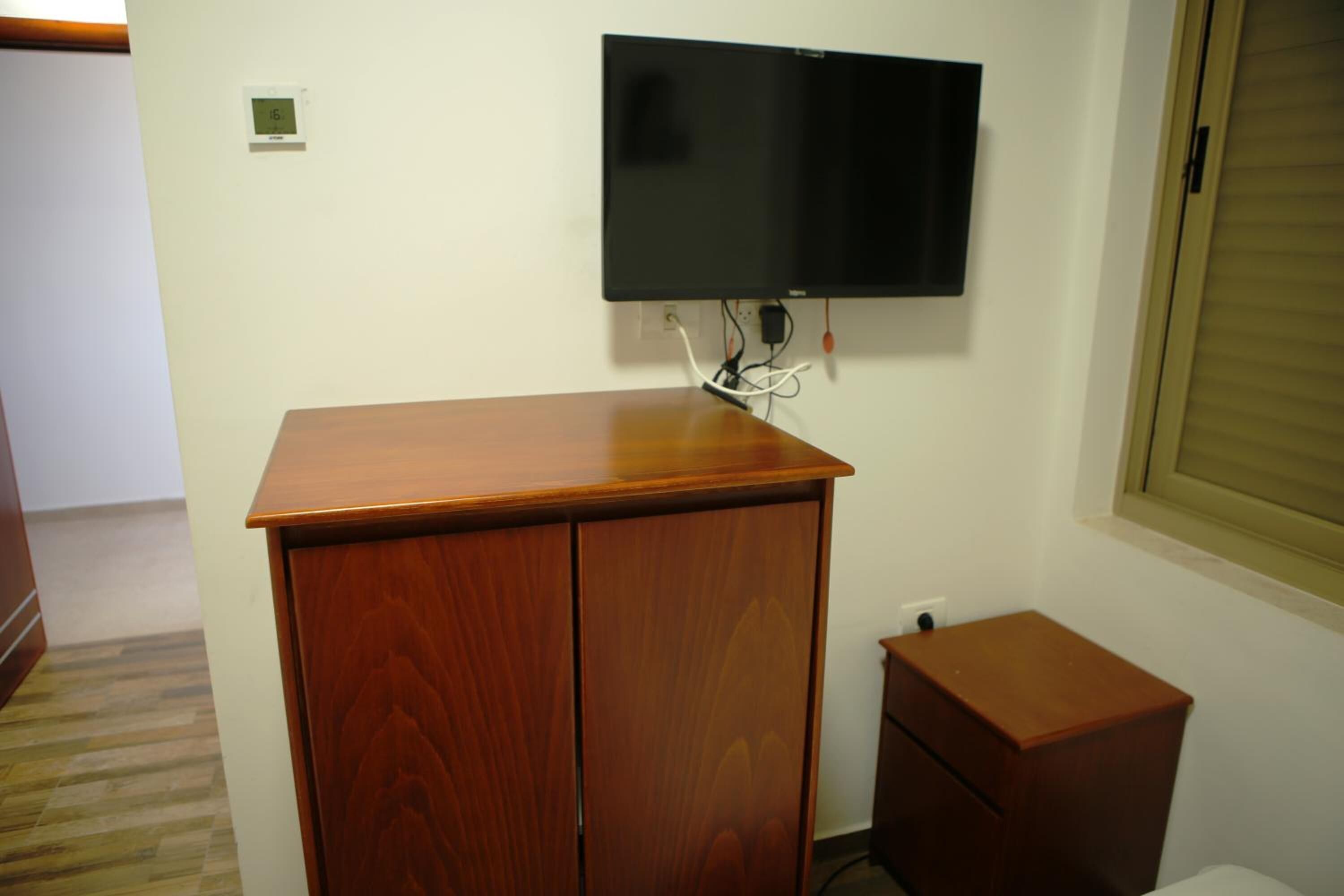 Una habitación con un armario de madera, un televisor montado en la pared y una mesita de noche de madera.