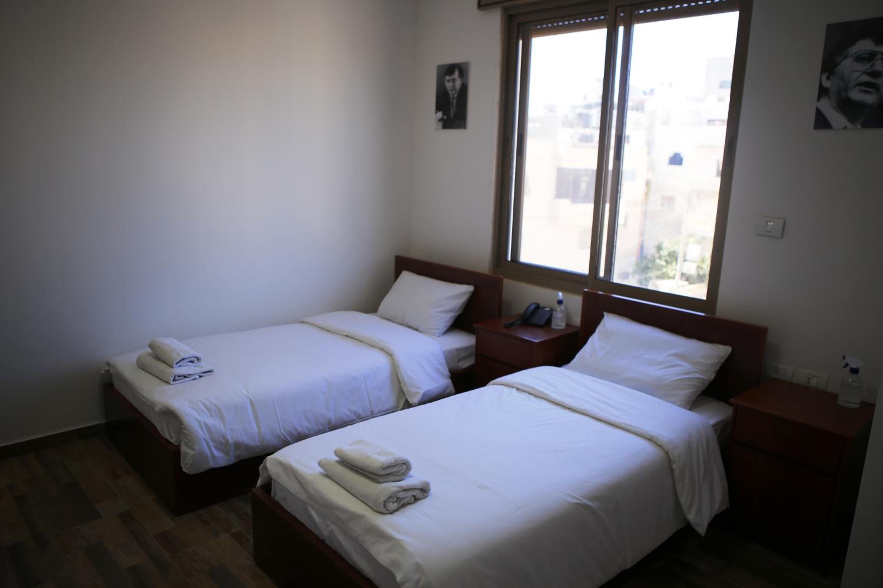 A hotel room with two single beds, a wooden headboard, a nightstand with a phone and a bottle, and a window with a city view.