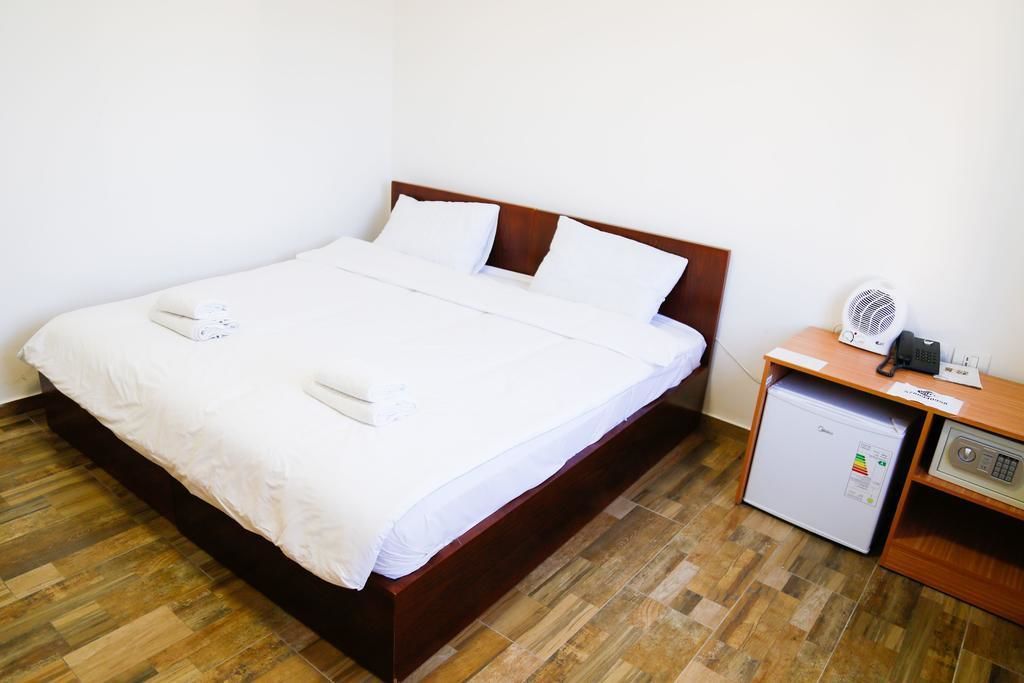 A neatly made bed with white linens and pillows, a small bedside table with a fan and a phone, and a compact refrigerator.