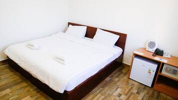 A neatly made bed with white linens and pillows, a small bedside table with a fan and a phone, and a compact refrigerator.