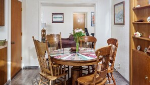 Zona pranzo con tavolo rotondo, sei sedie in legno e un vaso con fiori. A destra, un armadio in legno con oggetti decorativi.