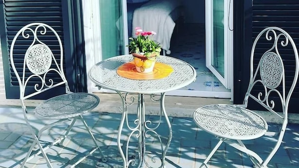 A patio with two metal chairs and a small round table, a potted plant, and a view into a room with a bed and a nightstand.