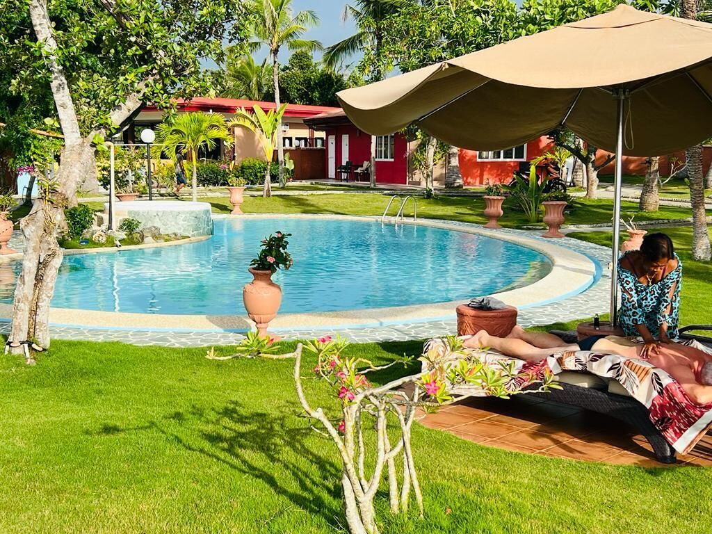 A pool area with a lounge chair and umbrella, surrounded by greenery and a small building in the background.