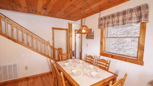 A dining area with a wooden table set for four, wooden chairs, a staircase, and a window with blinds. - Cozy 3BR Cabin w/ Long-Range Views, Hot Tub & Dog-Friendly (Todd)