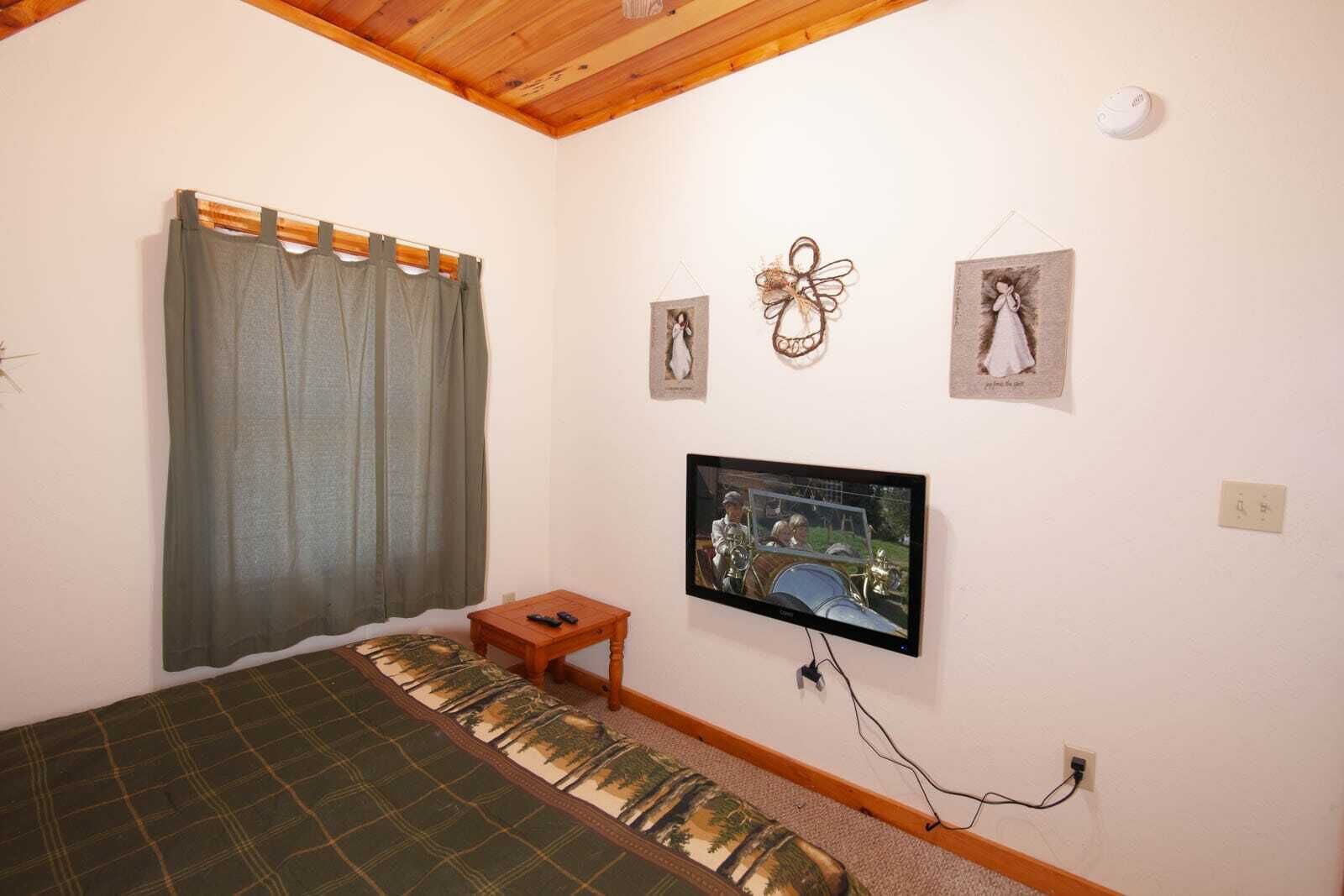 Ein Schlafzimmer mit einem Bett, einem an der Wand befestigten Fernseher, einem kleinen Nachttisch und einem Fenster mit Vorhängen.