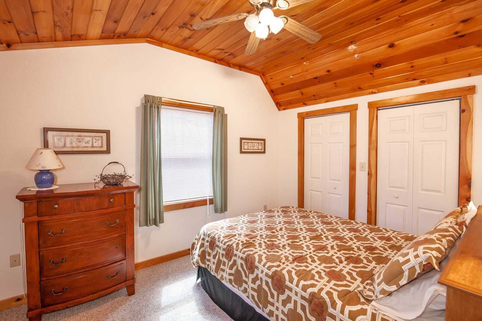 A bedroom with a wooden ceiling, a bed with a patterned bedspread, a dresser, a window with curtains, and two doors.