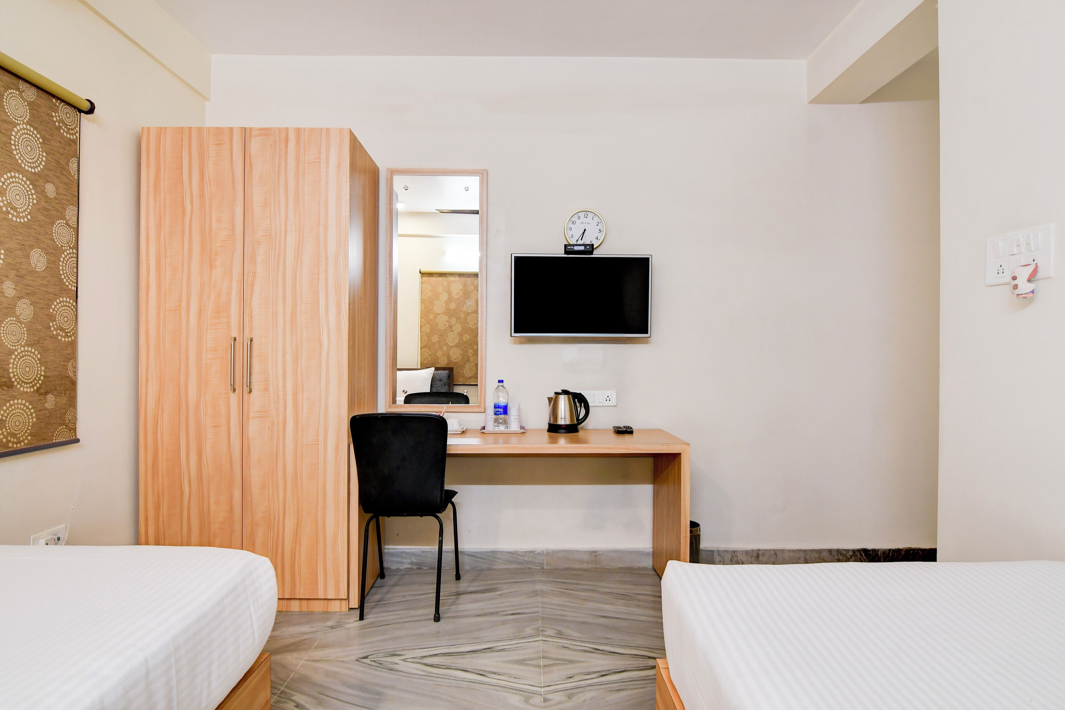 A hotel room with two beds, a wooden wardrobe, a desk with a chair, a flat-screen TV, a clock, and a kettle.