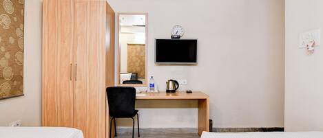 A hotel room with two beds, a wooden wardrobe, a desk with a chair, a flat-screen TV, a clock, and a kettle.