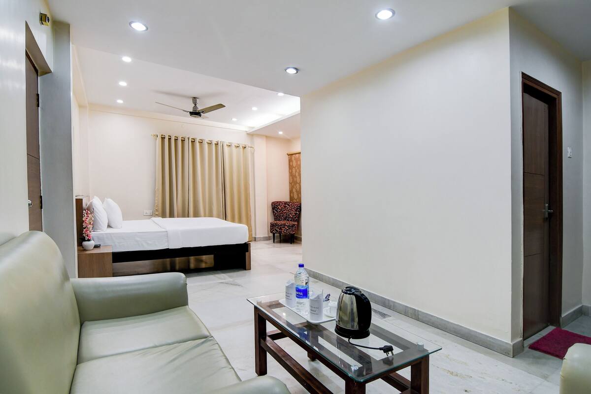 A hotel room with a bed, a sofa, a coffee table with a kettle, a water bottle, and a chair.