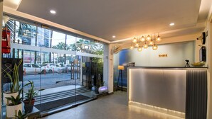 A modern hotel reception area with a glass entrance, potted plants, a reception desk, and a view of parked cars outside. - The Inn (Kolkata)