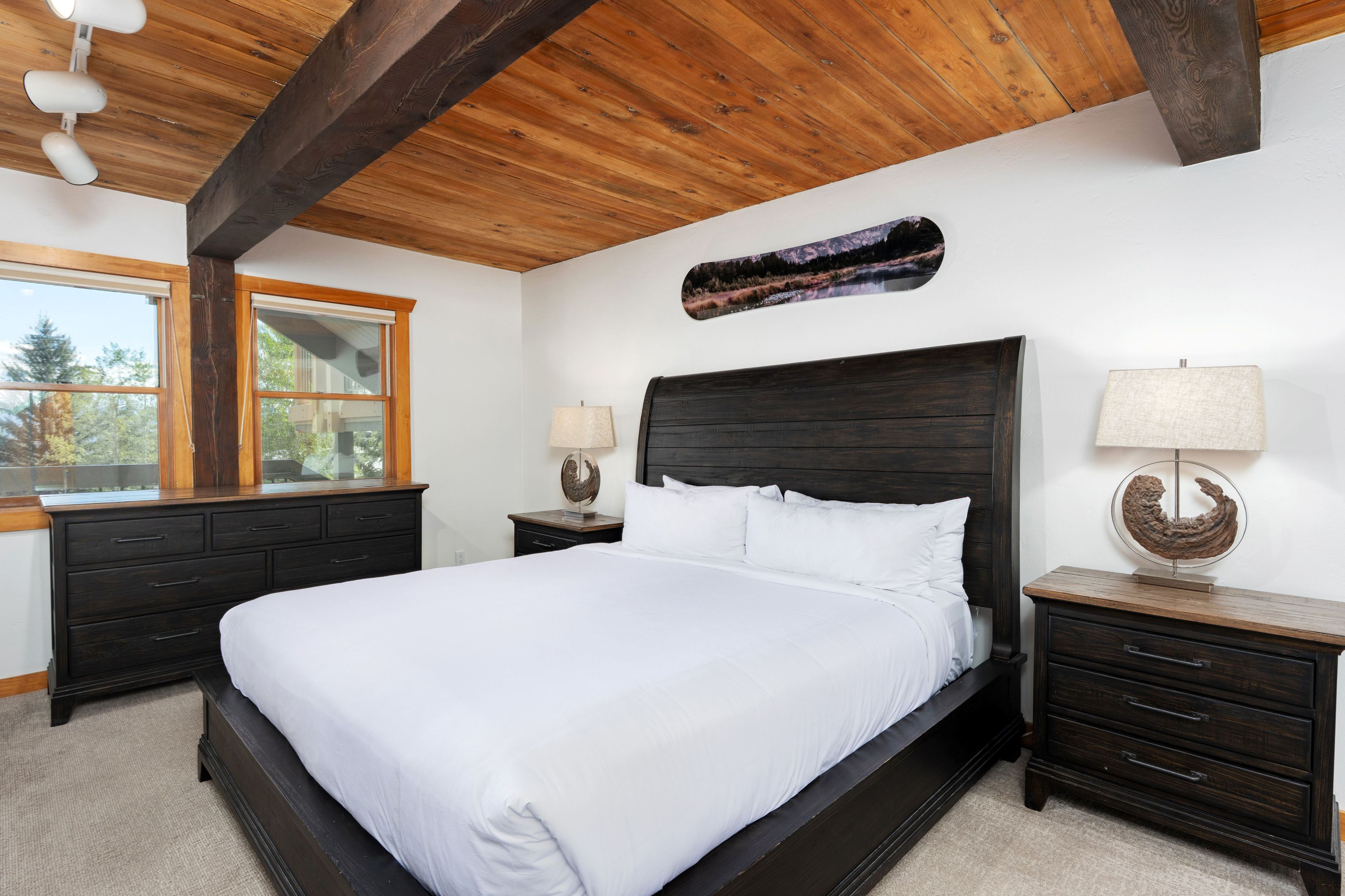 Une chambre à coucher avec un grand lit, une tête de lit en bois, deux tables de chevet avec des lampes, une fenêtre et une œuvre d’art accrochée au mur.