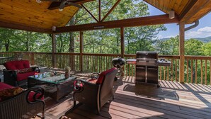 En overdækket terrasse med udendørs møbler, grill og udsigt til træer.