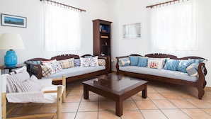 Ein Wohnzimmer mit zwei Holzsofas, einem Couchtisch und einem Holzstuhl. Ein Bücherregal und eine Lampe auf einem Beistelltisch sind ebenfalls vorhanden.