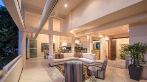 A spacious living area with a large sectional sofa, a small round table, and striped ottoman. There is a kitchen in the background, and the space is well-lit with natural light.