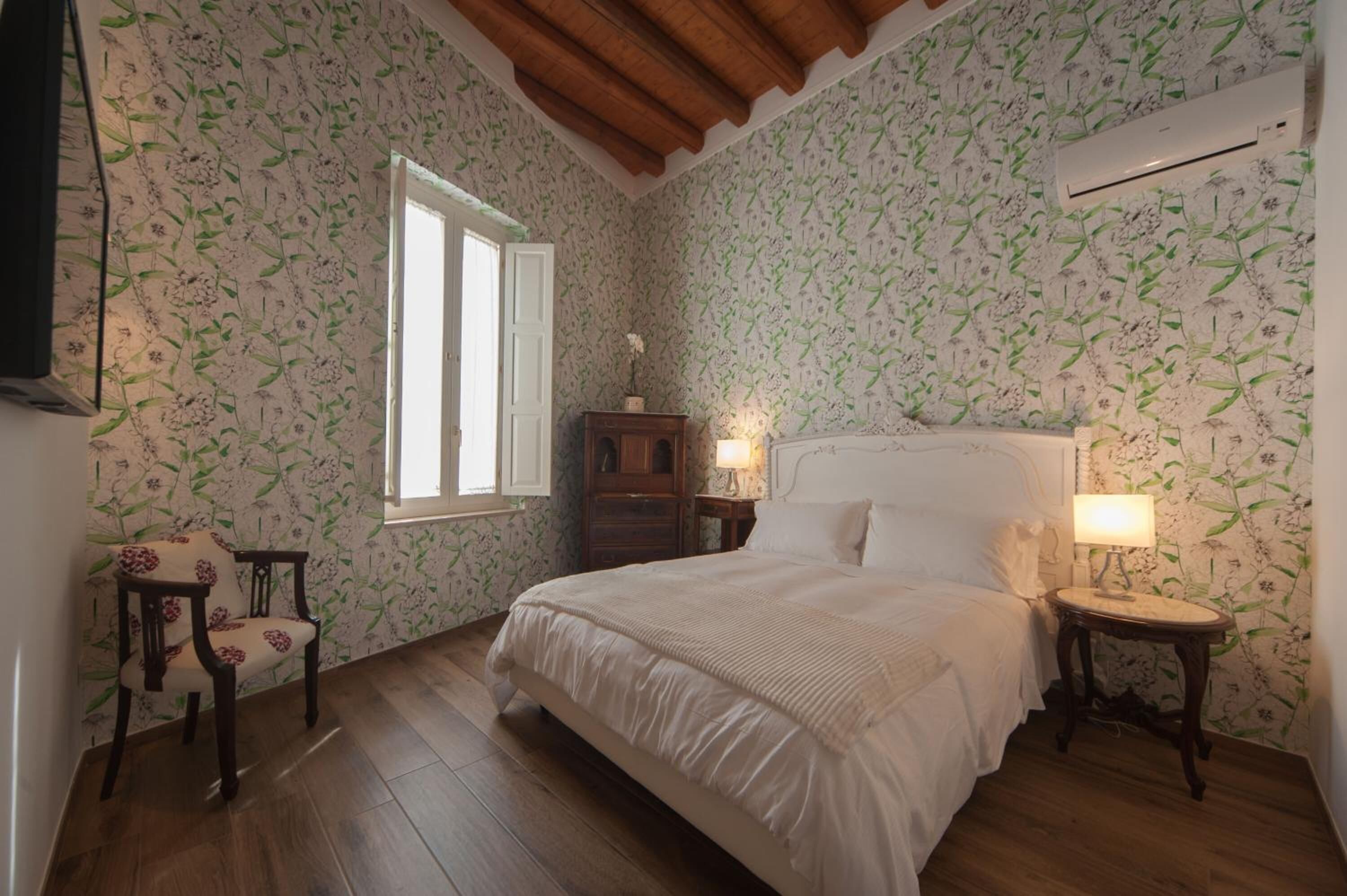 A bedroom with floral wallpaper, a wooden bed, a nightstand with a lamp, a chair, and a window.