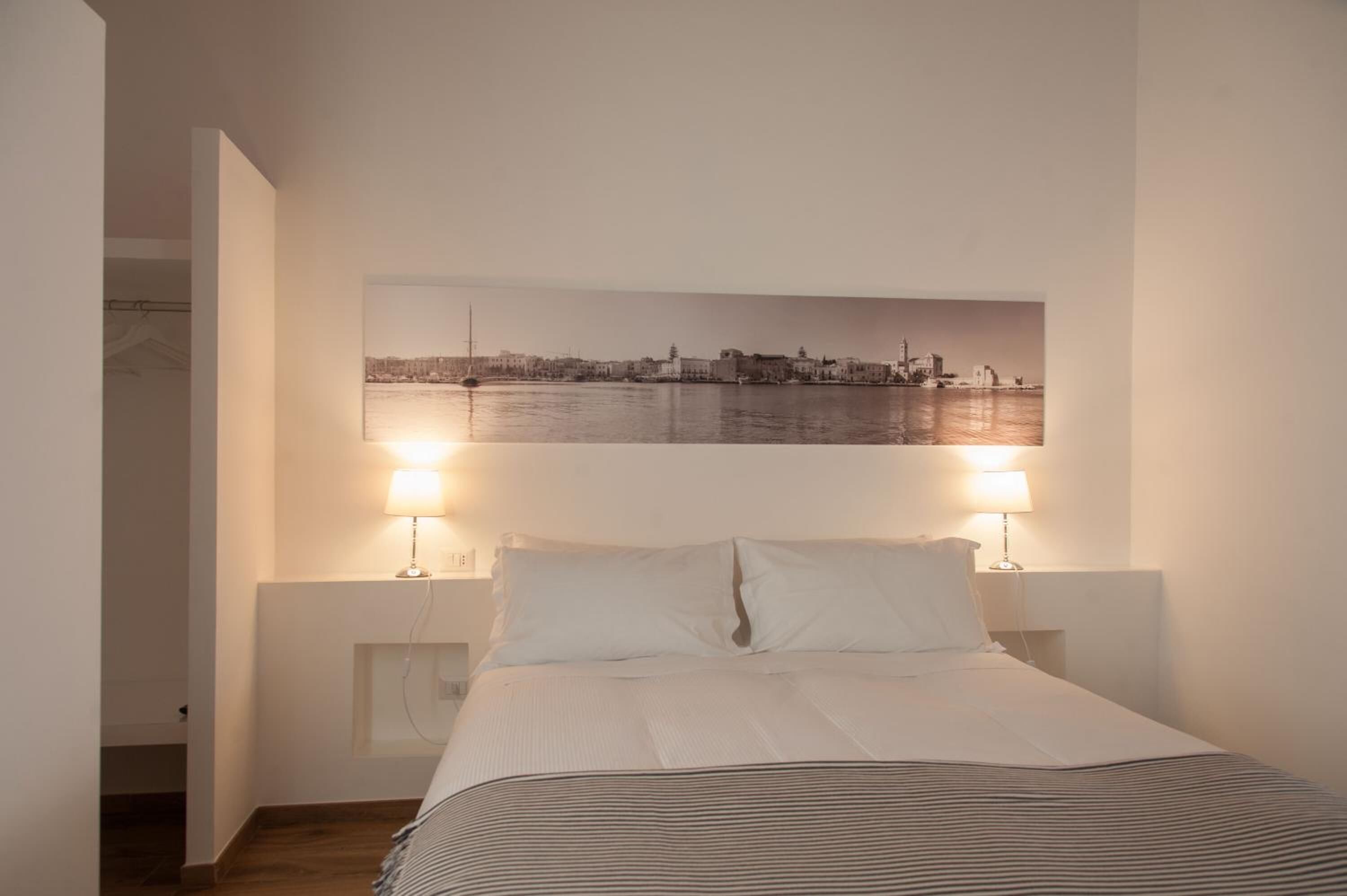 A bedroom with a bed, two bedside lamps, a cityscape painting on the wall, and a view of the water.