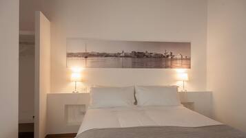 A bedroom with a bed, two bedside lamps, a cityscape painting on the wall, and a view of the water.