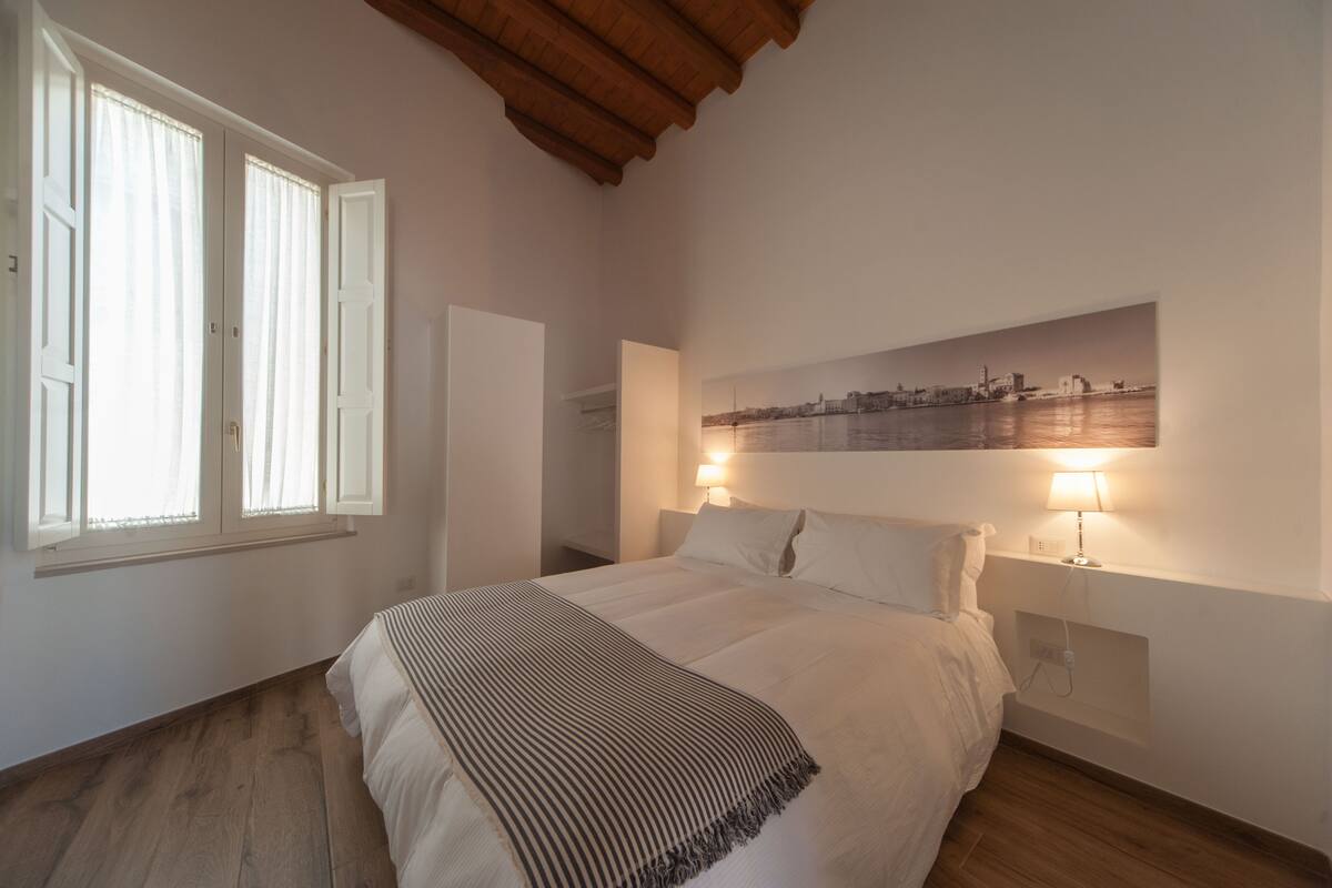 A bedroom with a bed, a cityscape painting, a window with curtains, and a wooden ceiling.