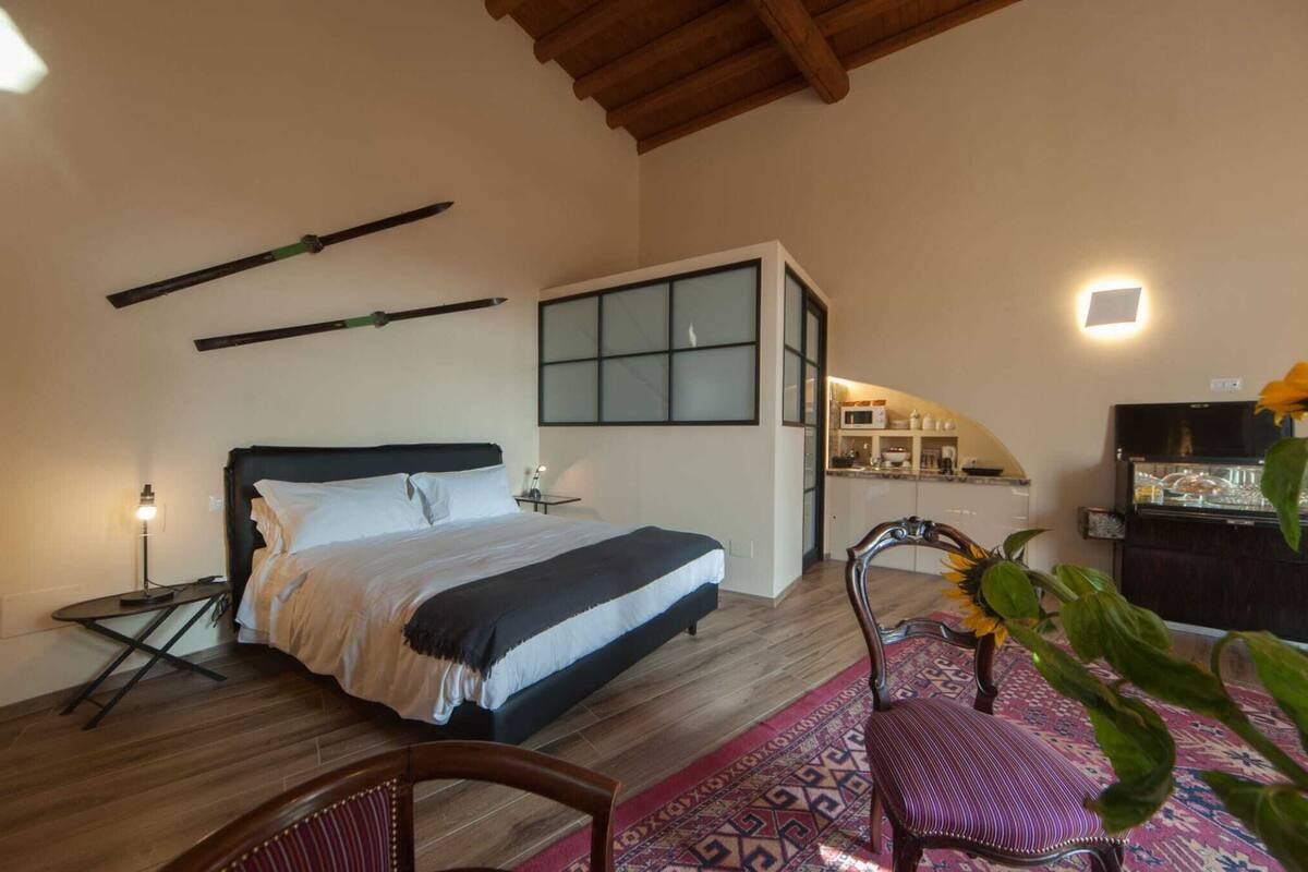 A bedroom with a bed, a nightstand, a chair, a window, and a view of a kitchen area.