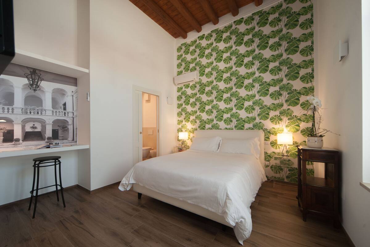 A bedroom with a bed, a window with a view of an architectural structure, a wall with a leafy pattern, and a small stool.