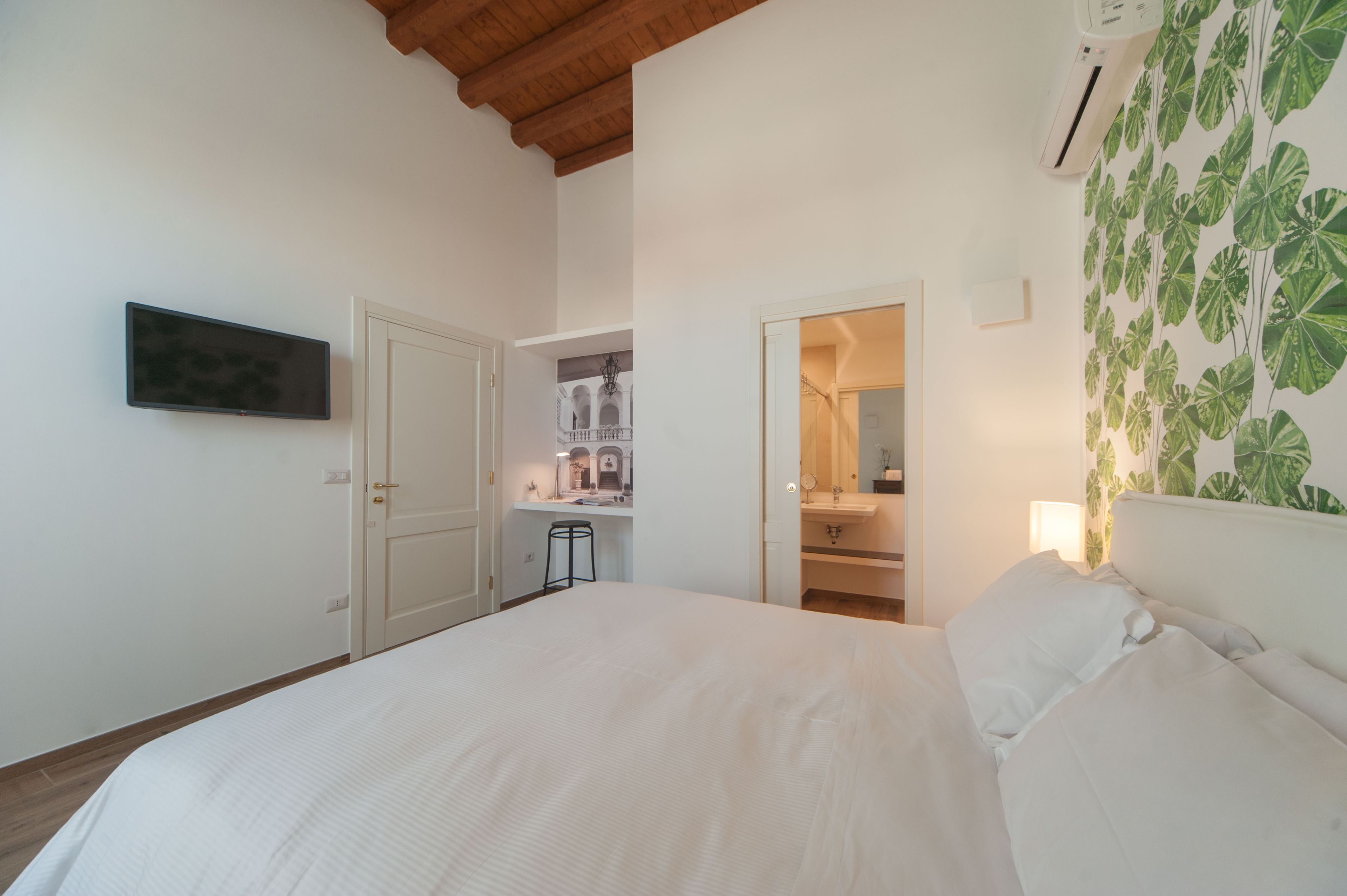 A bedroom with a large bed, a TV mounted on the wall, a desk with a chair, and a bathroom visible through an open door.