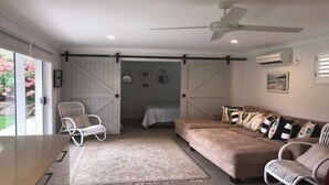 Un vaste espace de vie comprenant un grand canapé, un fauteuil blanc et une table à manger. On y trouve également un ventilateur de plafond, un lit avec vue et une fenêtre ornée de fleurs roses.