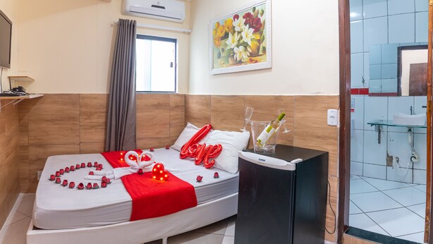 Una habitación de hotel con cama, una mesita de noche con una botella y un vaso, un cuadro en la pared y un baño visible a través de una puerta abierta.