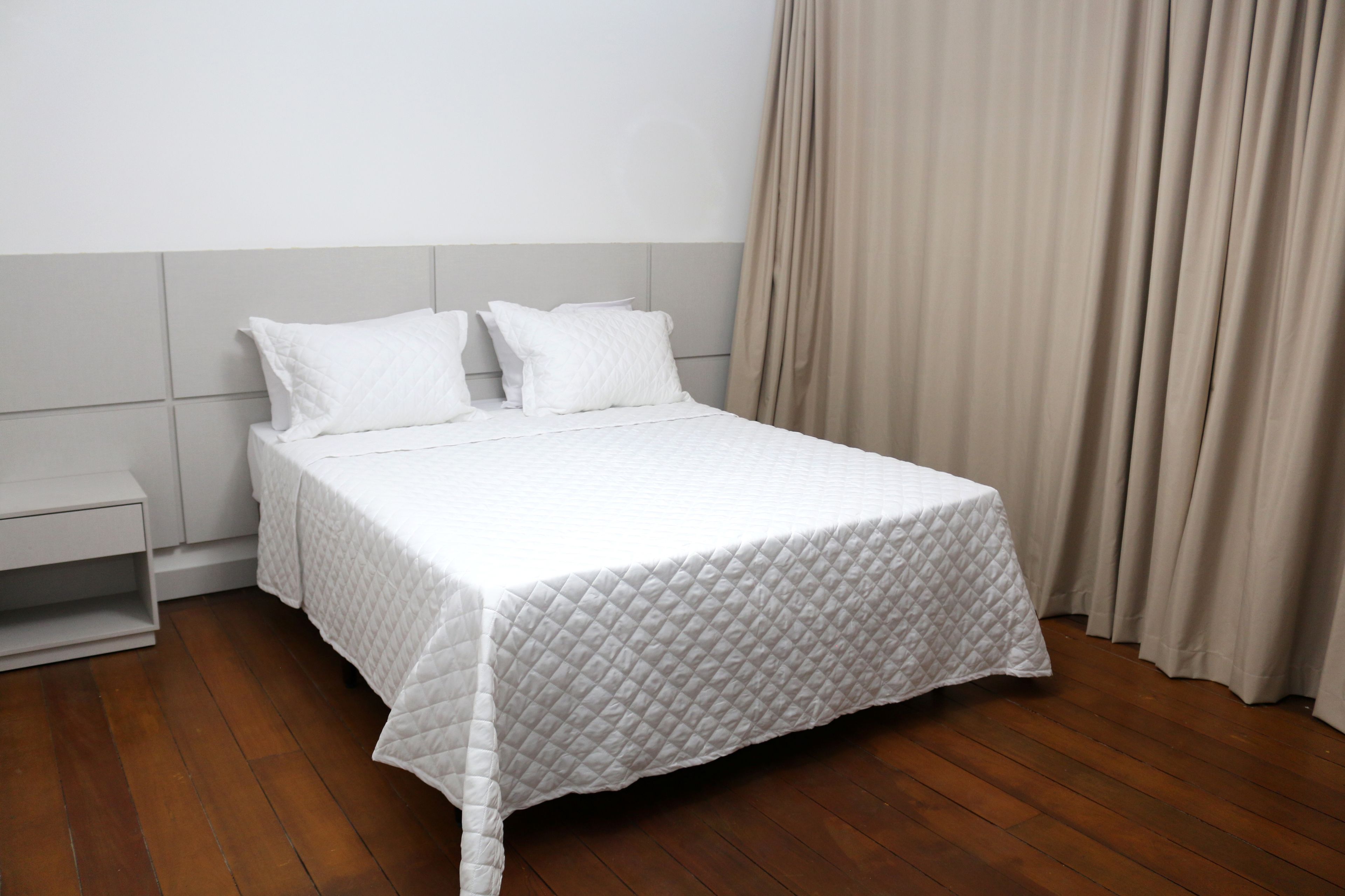 A neatly made bed with a white quilt, a wooden nightstand, and a beige curtain.