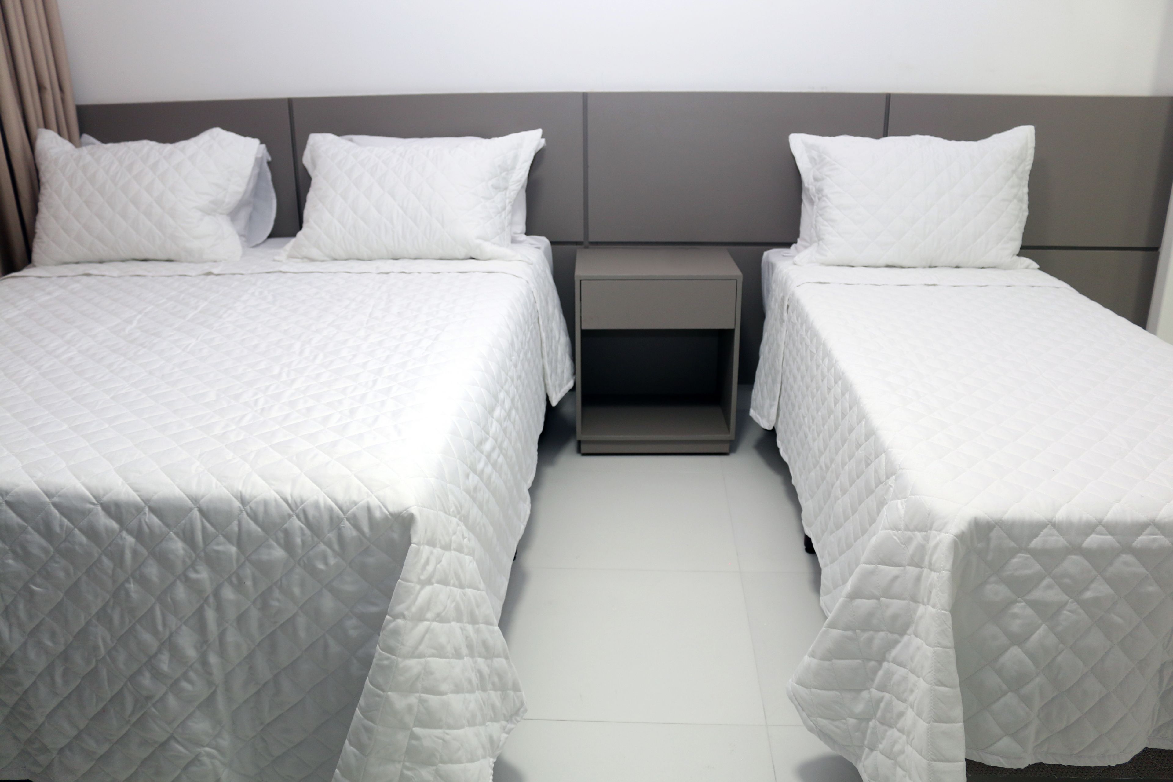 Quarto de hotel com duas camas de solteiro, cada uma com edredom branco acolchoado e almofadas a combinar, uma pequena mesa de cabeceira e cabeceira de cor neutra.
