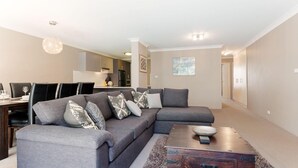 A modern living room with a sectional sofa, dining table, and kitchen in the background. - Bay Parklands, 34 2 Gowrie Avenue - Air con, Wifi, Views, Pool, Spa & Tennis Court (Nelson Bay)
