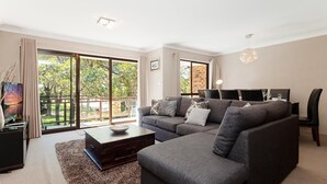 A modern living room with a grey sofa, a coffee table, and a TV. There are large windows with a view of greenery outside. - Bay Parklands, 34 2 Gowrie Avenue - Air con, Wifi, Views, Pool, Spa & Tennis Court (Nelson Bay)