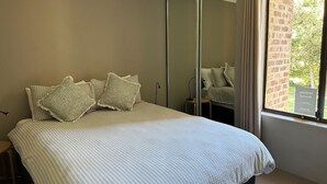 A bedroom with a bed, pillows, a mirror, and a window with curtains. - Bay Parklands, 34 2 Gowrie Avenue - Air con, Wifi, Views, Pool, Spa & Tennis Court (Nelson Bay)
