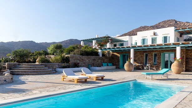 Zona de piscina con sillones de descanso, un muro de piedra y un edificio con paisaje montañoso.