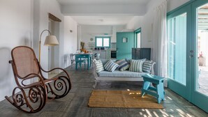 Amplia sala de estar con una mecedora, un sofá con cojines a rayas y una pequeña mesa. Se observa una cocina al fondo con armarios color verde azulado.