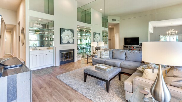 Una sala de estar moderna con un sofá gris, una mesa de centro y un televisor. Se observa un aparador de vidrio con objetos decorativos y una chimenea.