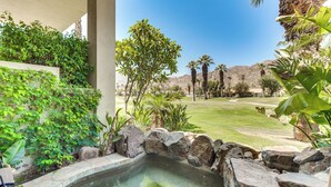 Un área de piscina con borde de piedra, abundante vegetación y palmeras al fondo.