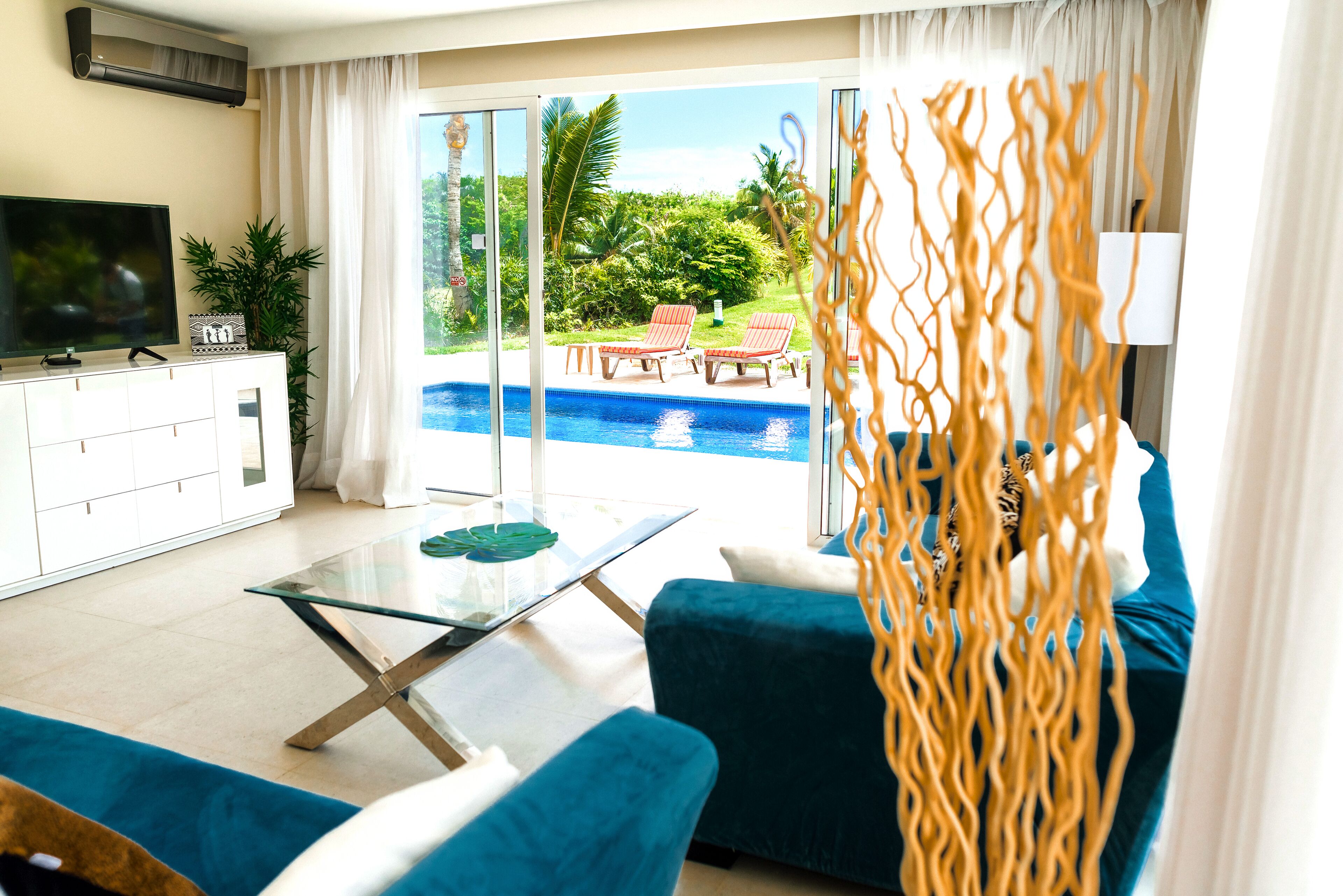 A living room with a glass coffee table, blue sofa, and a TV on a white cabinet. A glass door leads to a pool area with red lounge chairs.
