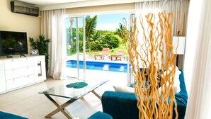 A living room with a glass coffee table, blue sofa, and a TV on a white cabinet. A glass door leads to a pool area with red lounge chairs. - Villa Fortuna Iberostar, private pool,  Bavaro beach, golf club, Wi-Fi (bavaro)